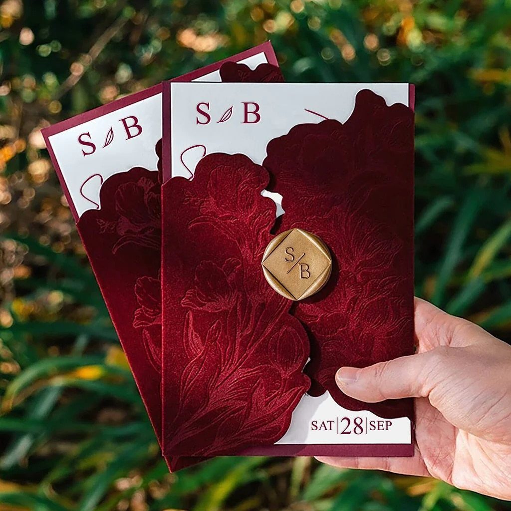 Burgundy floral laser-cut wedding invitations with a gold wax seal held against greenery.