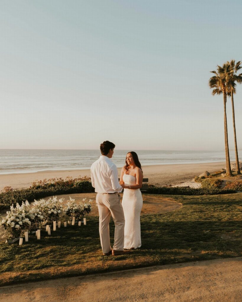 Couple standing together by white floral arrangements on a cliff overlooking the beach