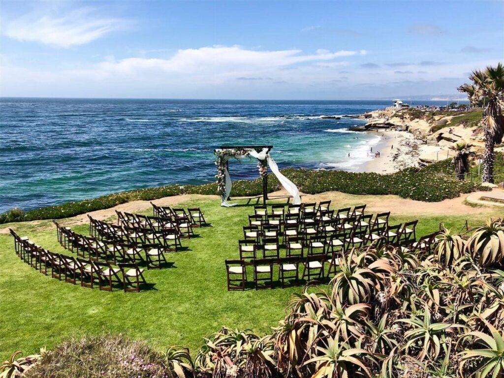 Wedding ceremony setup with wooden chairs and floral arch overlooking a sunny beach.