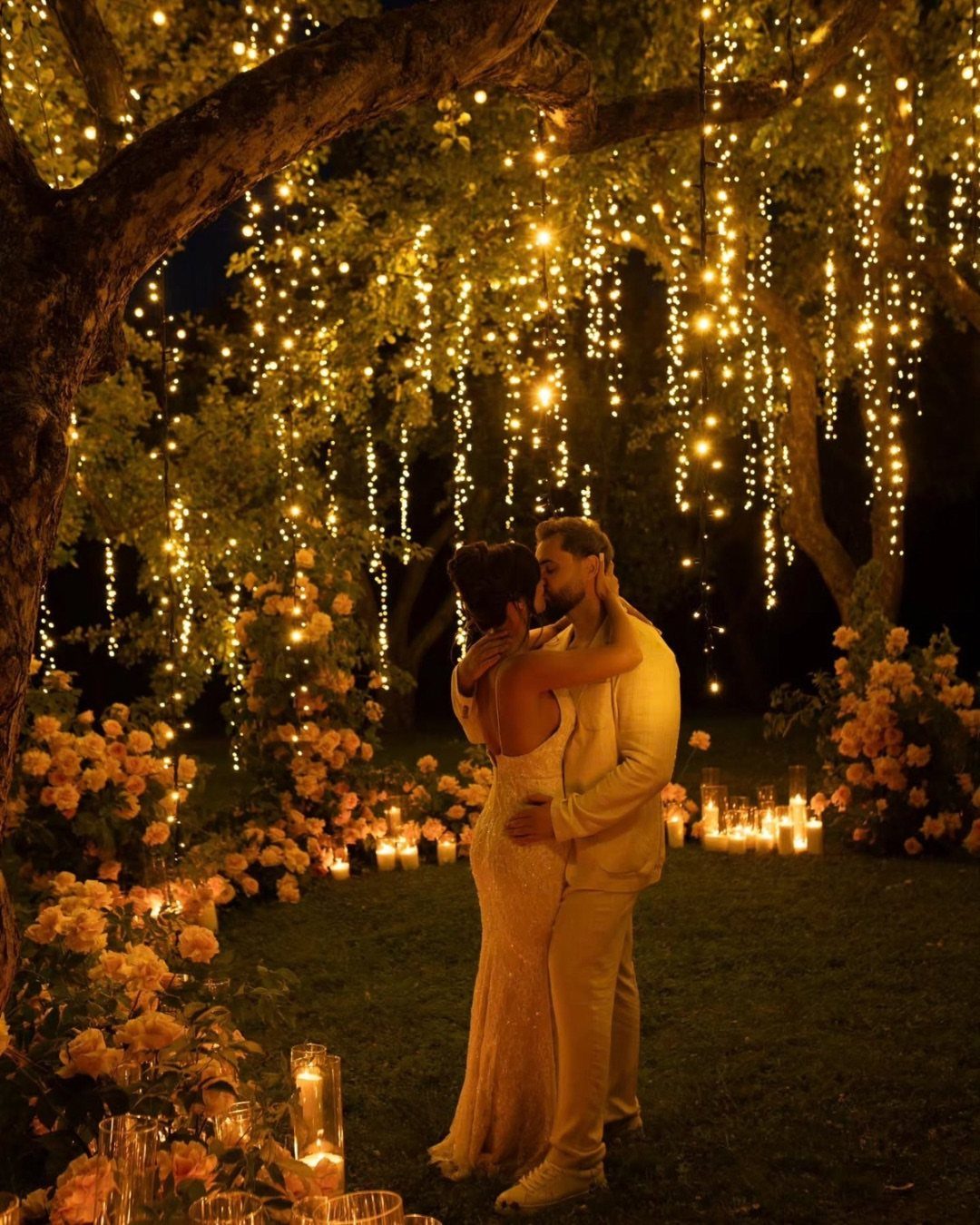 Wedding couple embracing under a tree decorated with fairy lights and peach roses.