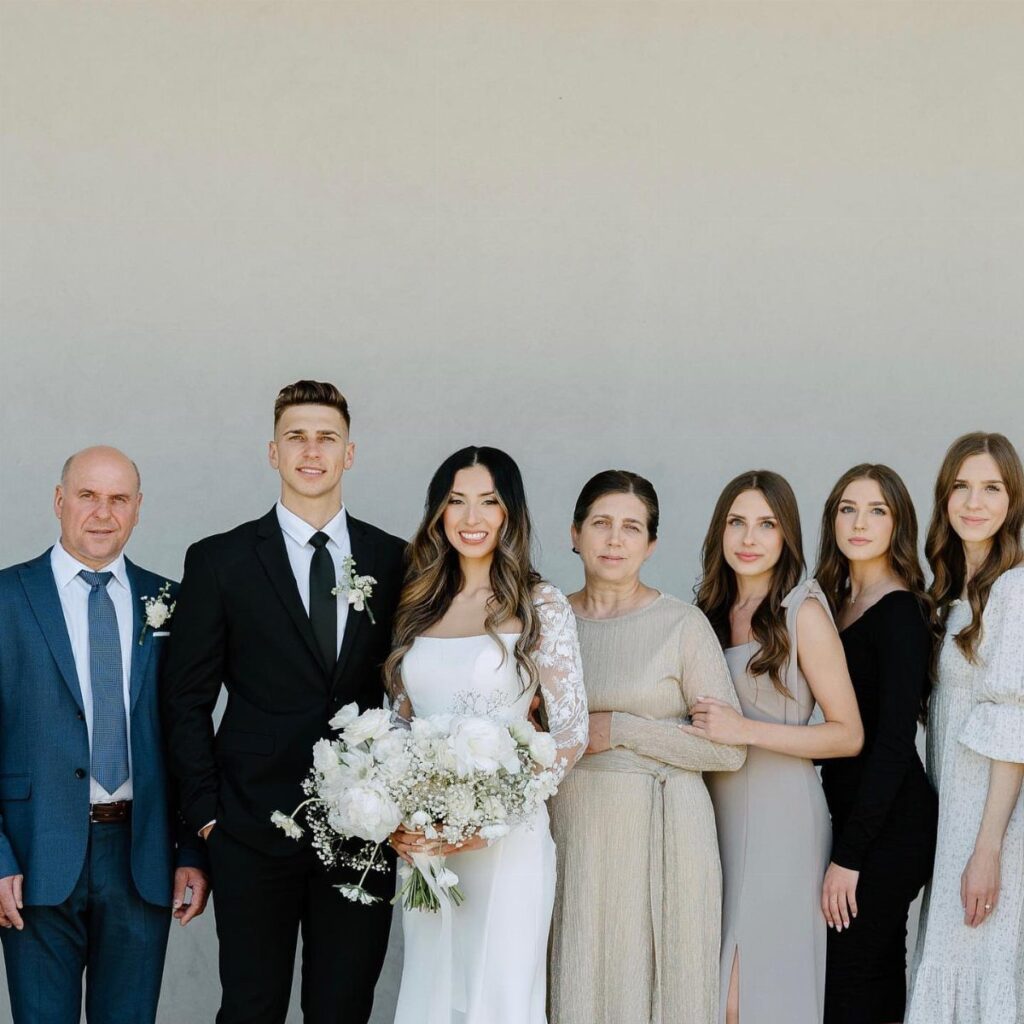 Bride and groom posing with five family members against a plain gray wall.