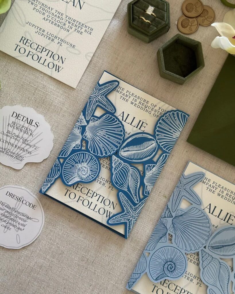 Blue laser-cut wedding invitations with seashell patterns resting on a linen surface.