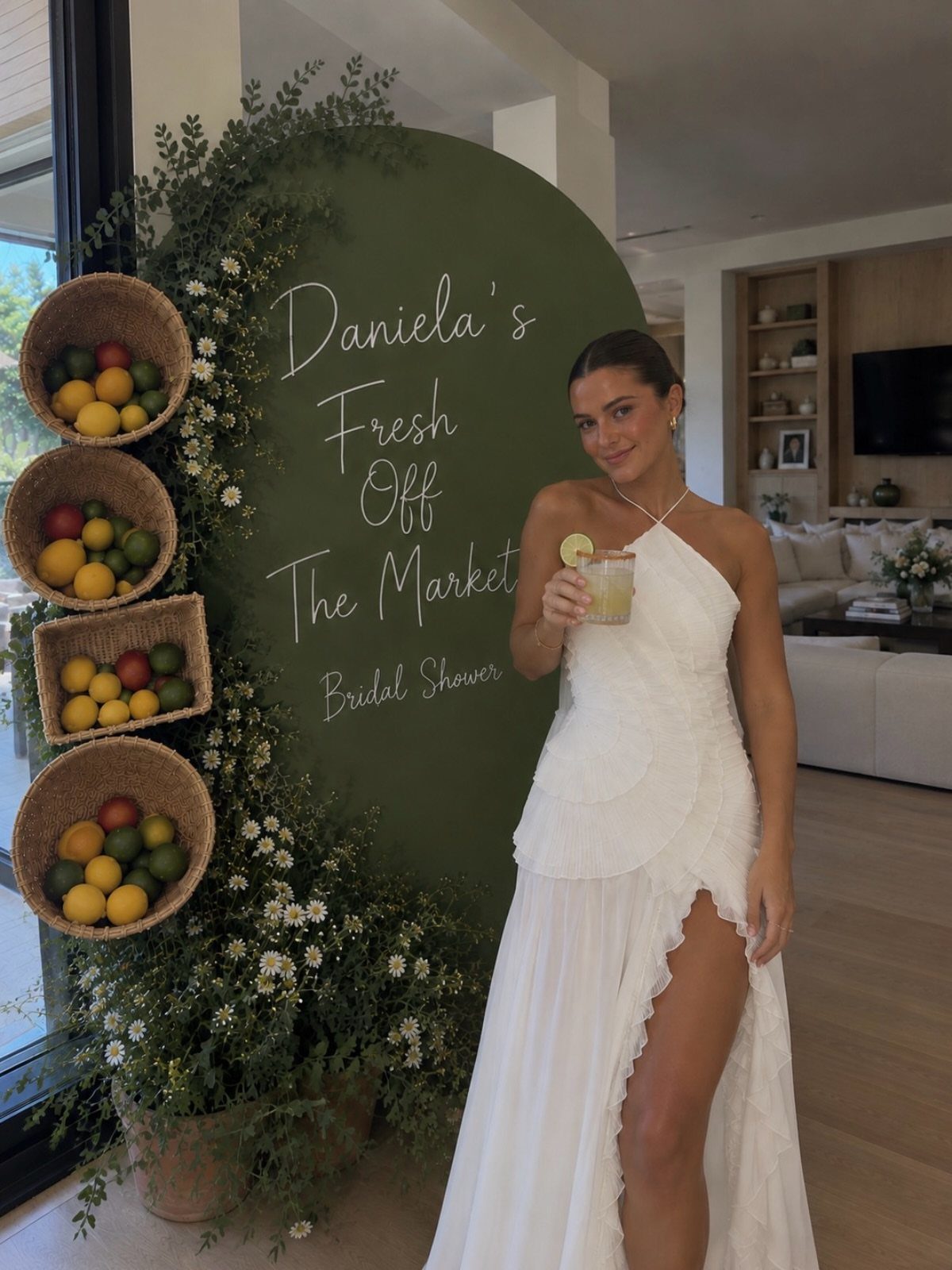 Woman in white dress posing by a green market themed bridal shower backdrop