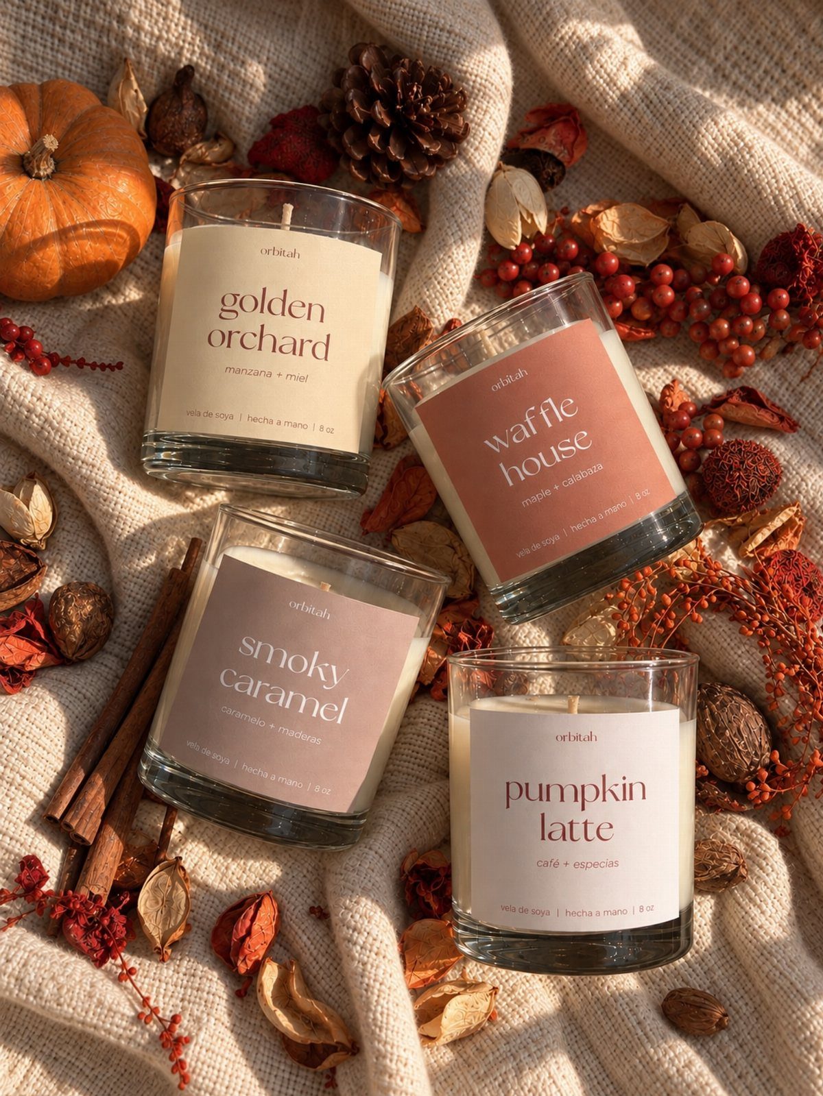 Four autumn-themed scented candles arranged on beige fabric with pumpkins and pinecones.
