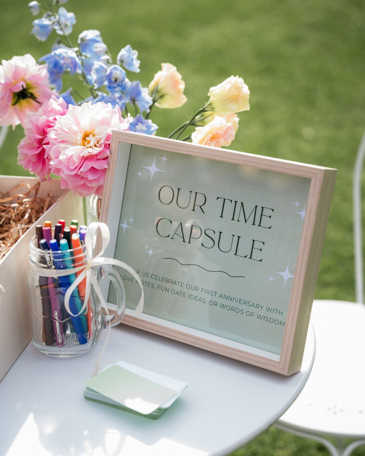 Time capsule station with a framed sign, colorful markers, and fresh flowers outdoors.