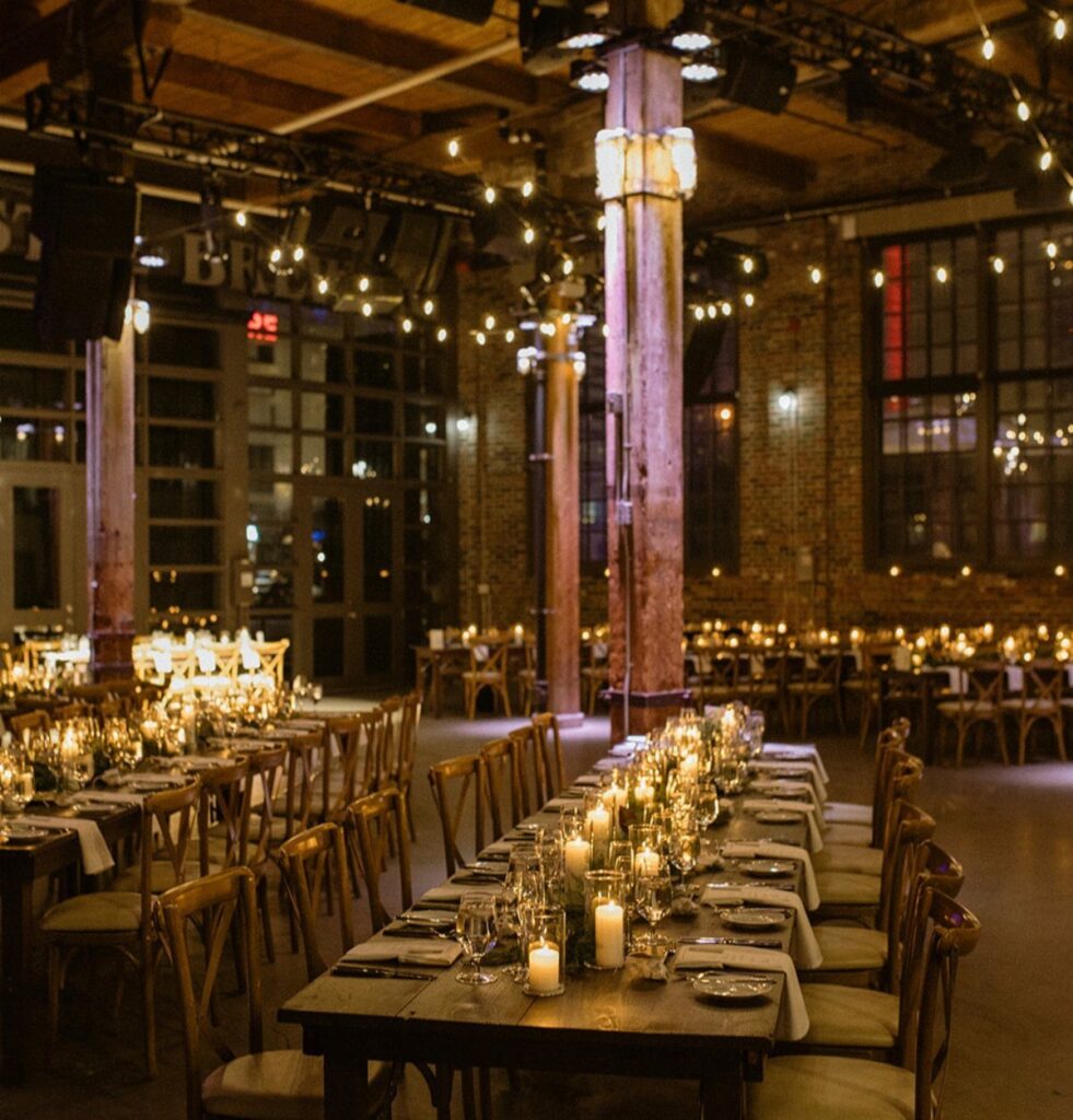 Industrial venue with long wooden tables, white candles, and warm string lighting.