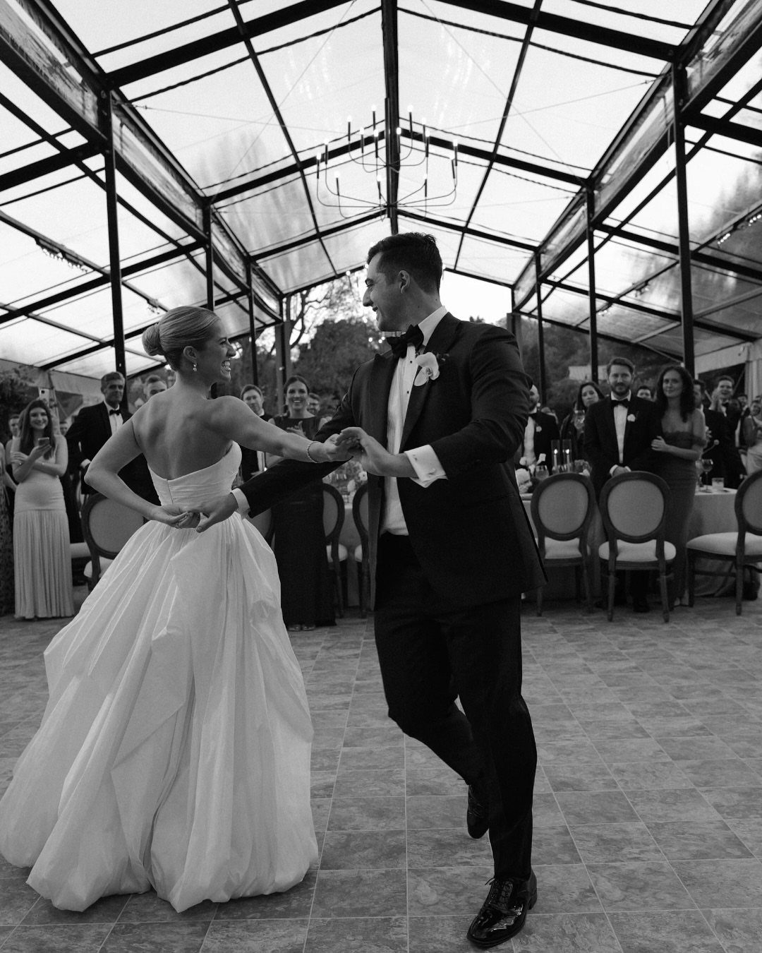 Bride and groom dancing in a glass pavilion wedding venue with guests watching.