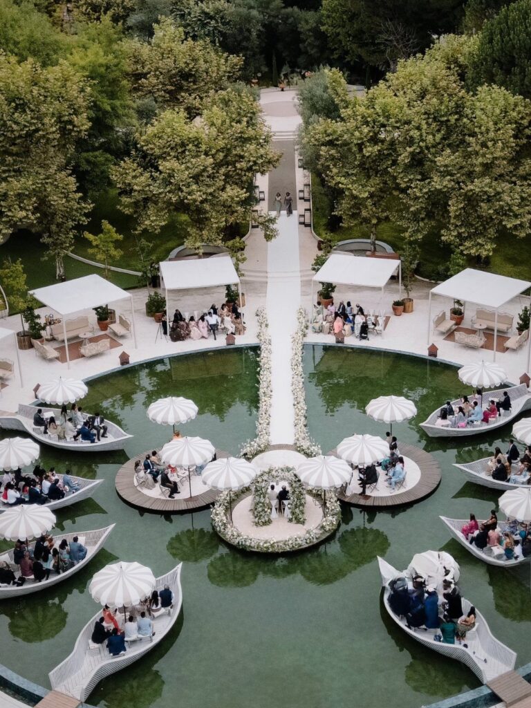 Wedding ceremony setup with boat-shaped seating and floral aisle over a green pond.